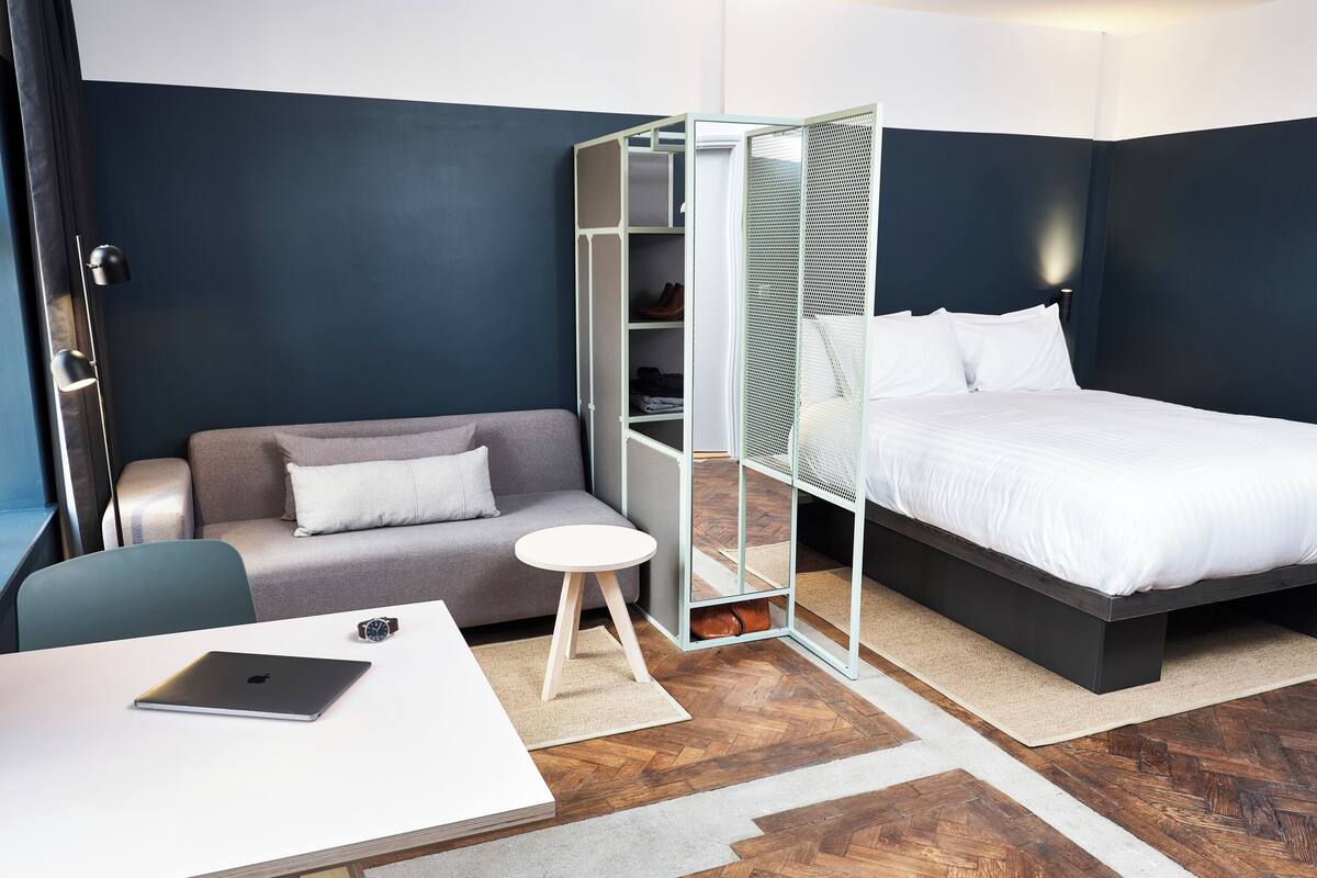 Ein modernes Hotelzimmer mit grauem Sofa, einem kleinen runden Tisch mit einem Laptop, einem Bett mit weißer Bettwäsche und einem begehbaren Kleiderschrank mit Maschendrahtür.