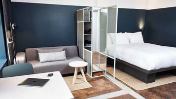 Una habitación de hotel moderna con un sofá gris, una pequeña mesa redonda con una computadora portátil, una cama con ropa blanca y un vestidor con puerta de malla.