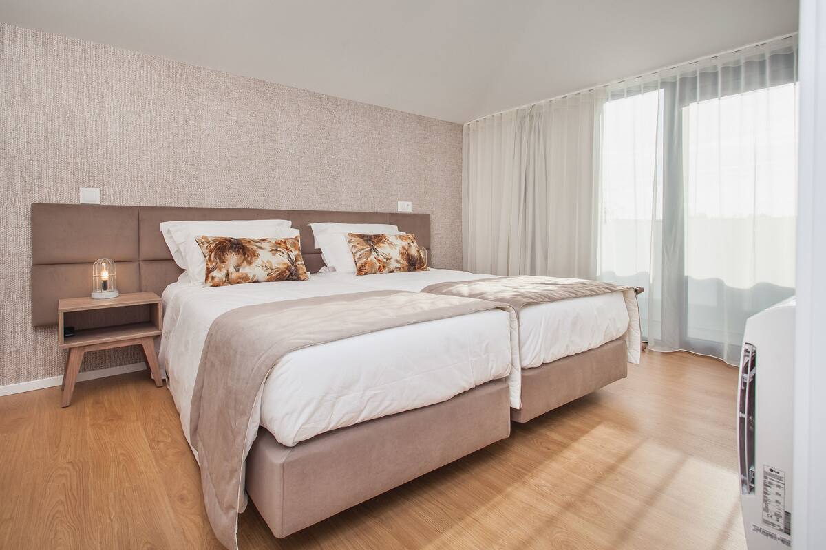 A modern hotel room with a large bed, a bedside table with a lamp, and a view through sheer curtains.