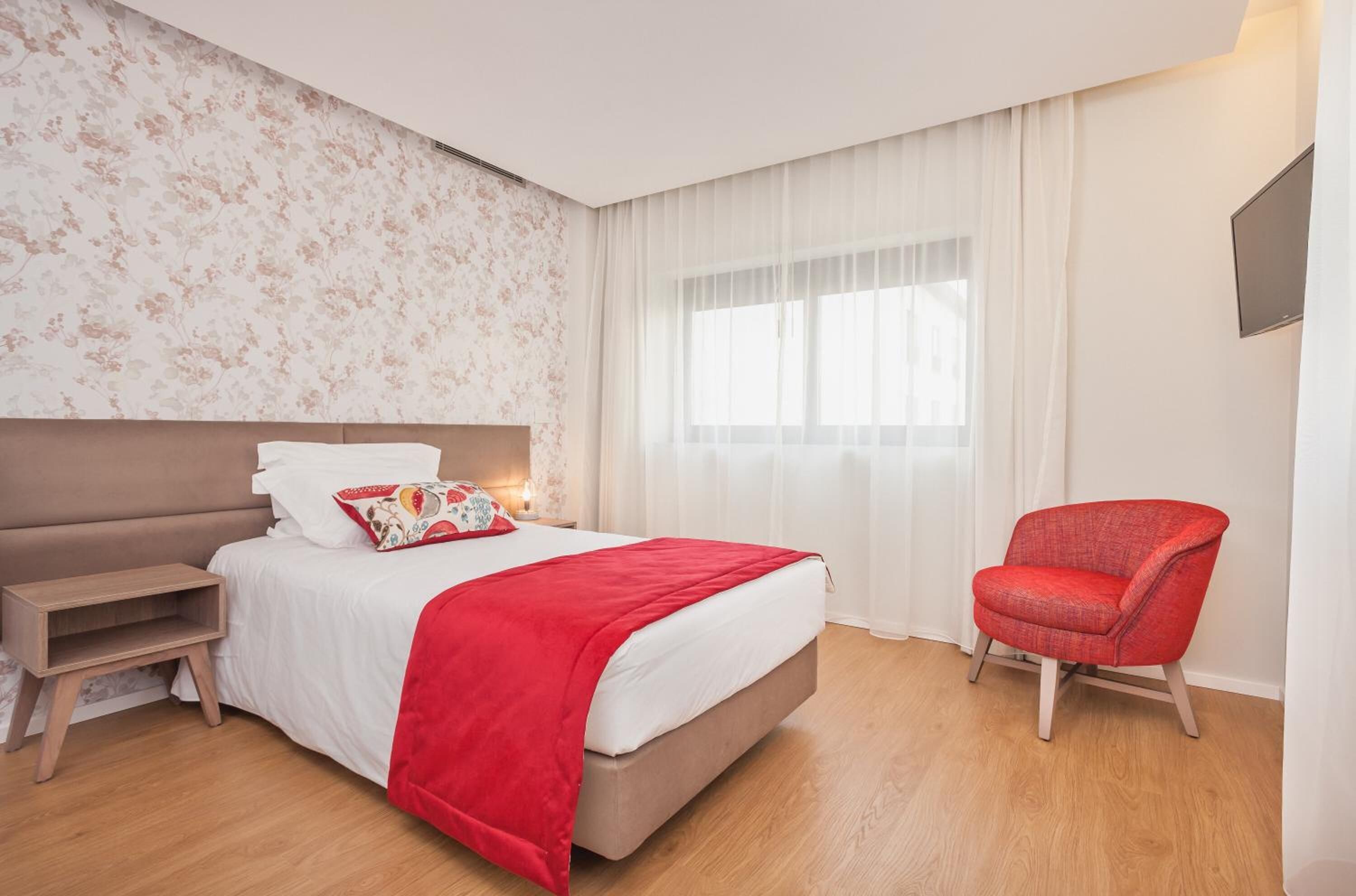Quarto de hotel com cama, mesa de cabeceira, uma cadeira e uma janela grande com cortinas.