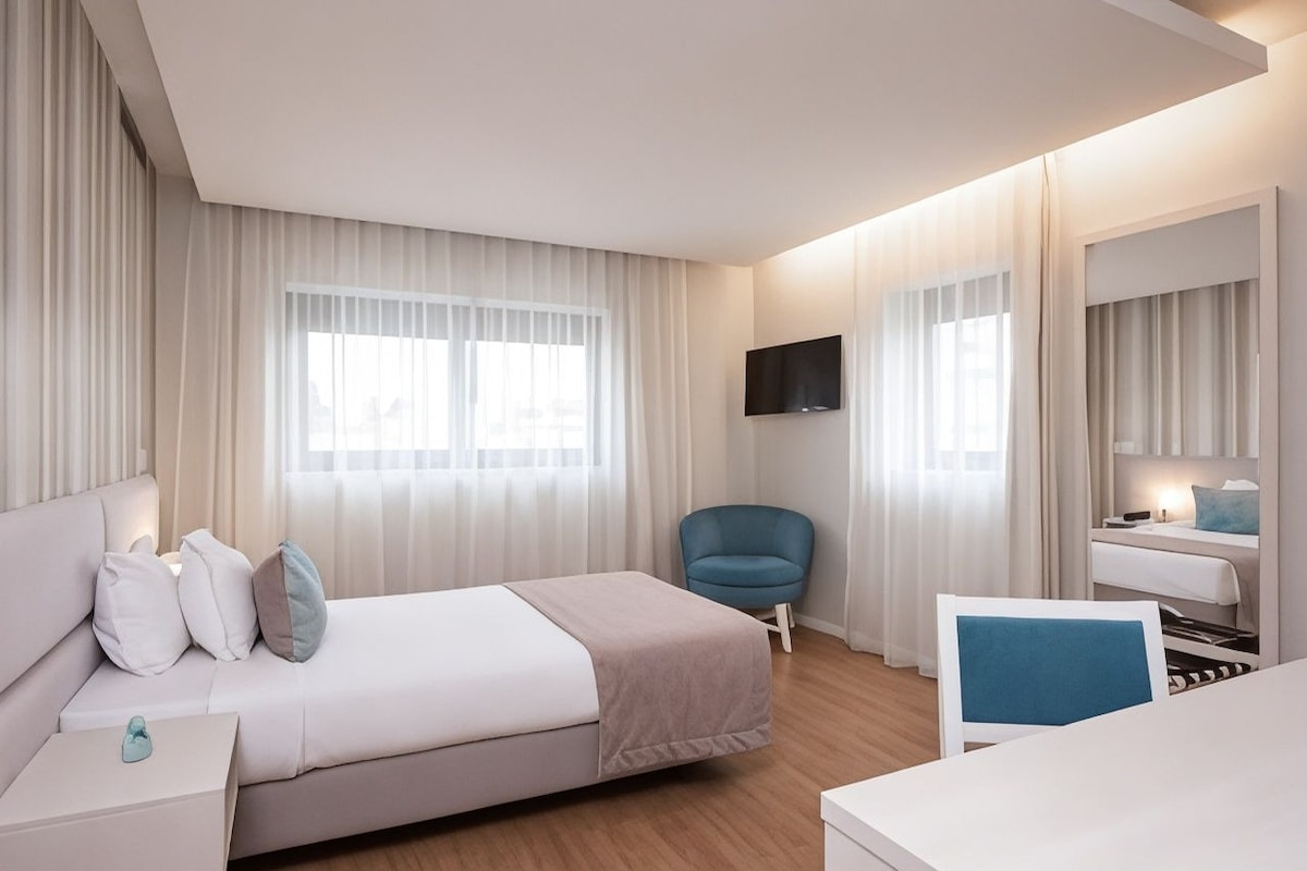 A hotel room with a bed, a desk, a chair, a TV, and a window with curtains.