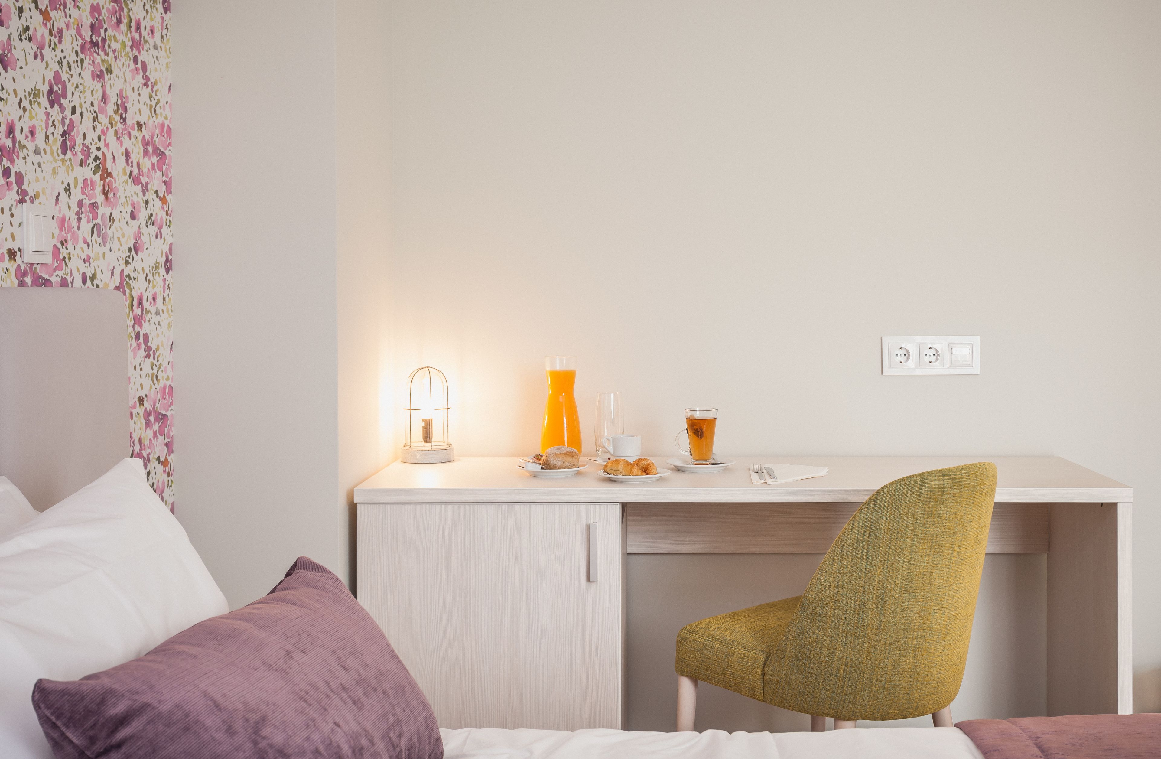 A neatly arranged bedroom with a bed, a desk with breakfast items, and a chair.
