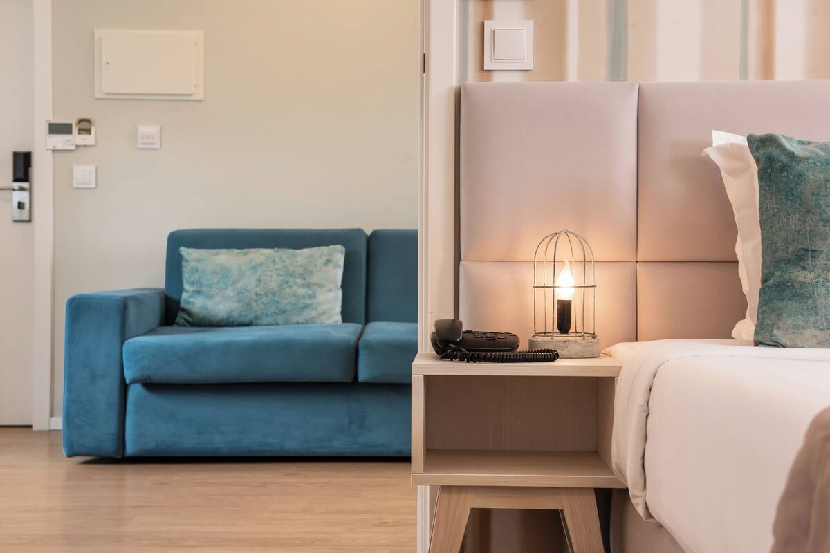 A modern hotel room with a blue sofa, a bedside table with a lamp, and a bed with white bedding.