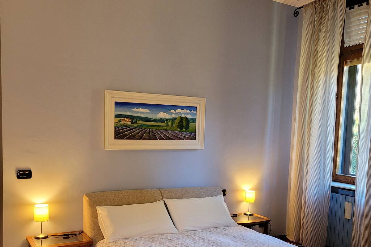A bedroom with a bed, two bedside tables with lamps, a framed landscape painting, and a window with curtains.