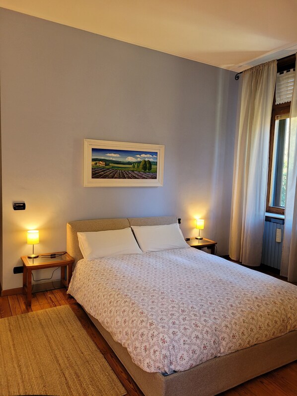 A bedroom with a bed, two bedside tables with lamps, a framed landscape painting, and a window with curtains.