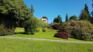 A well-maintained garden with a variety of trees and shrubs, a clear sky, and a building with a distinctive roof in the background. - Villa Tavallini (Pollone)