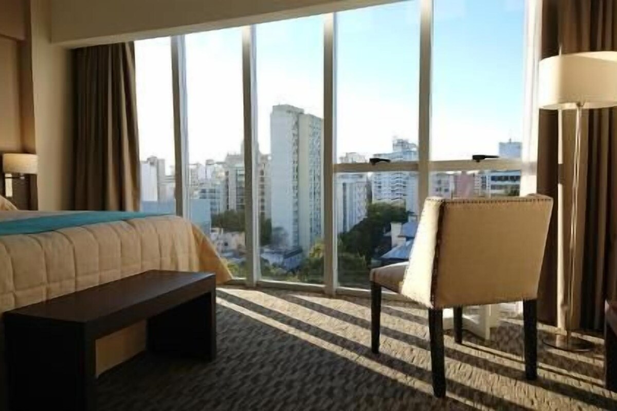 A hotel room with a large bed, a chair, a small table, and a view of the city through large windows.
