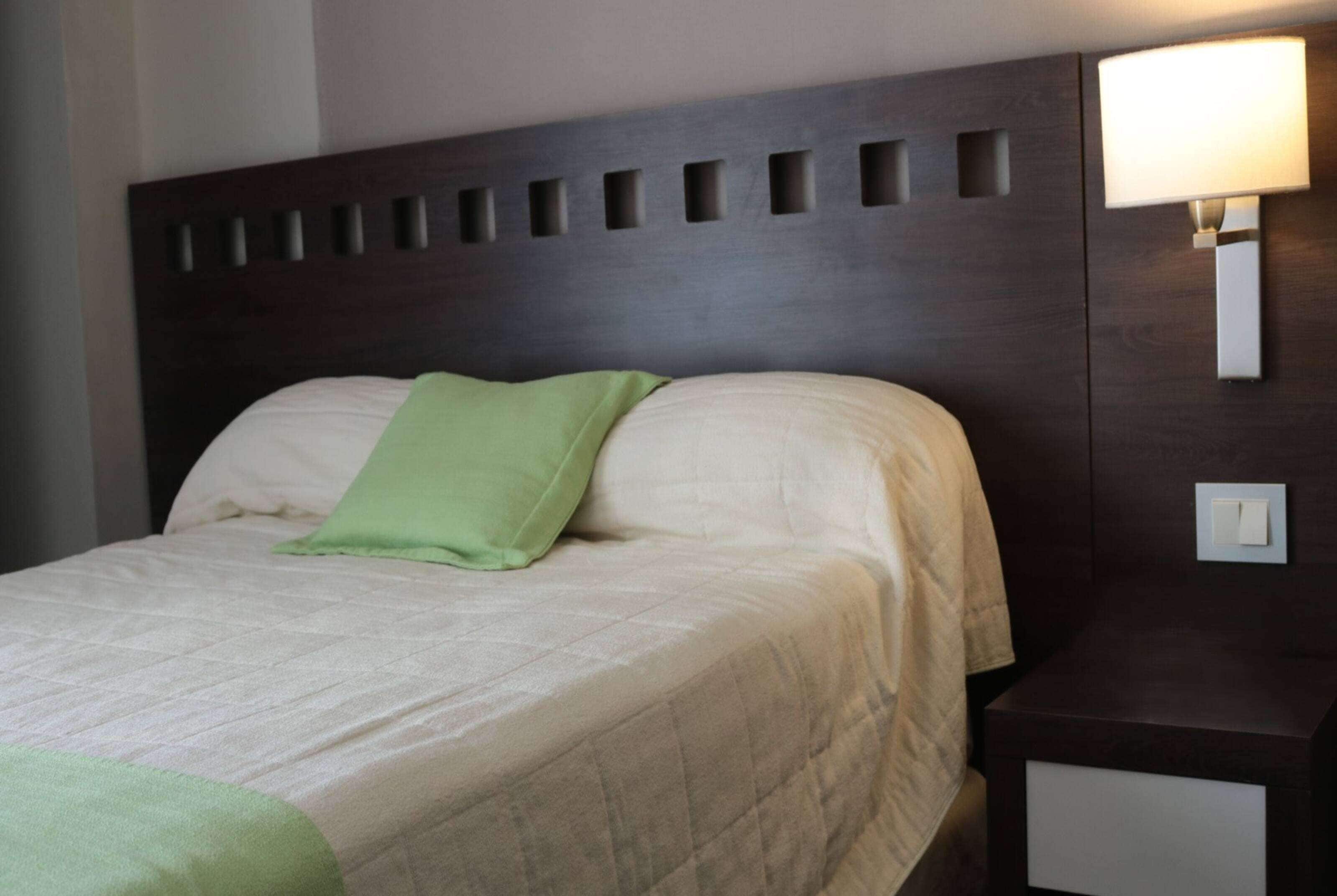 A neatly made bed with a green pillow and a white blanket, a wooden headboard with cut-out designs, and a bedside table with a lamp.