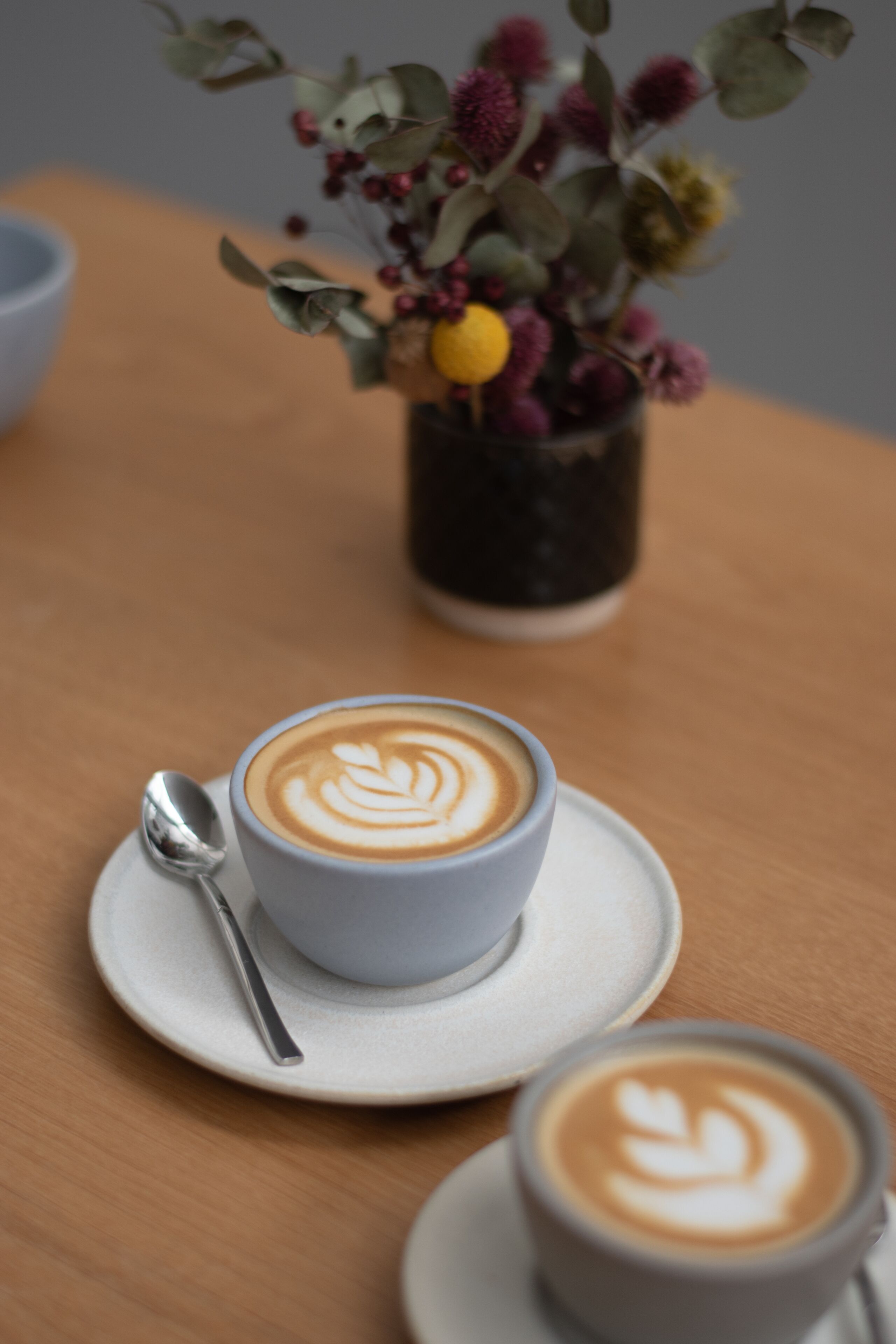 A cup of coffee with latte art on a saucer, a spoon, and a small potted plant with various flowers in the background.