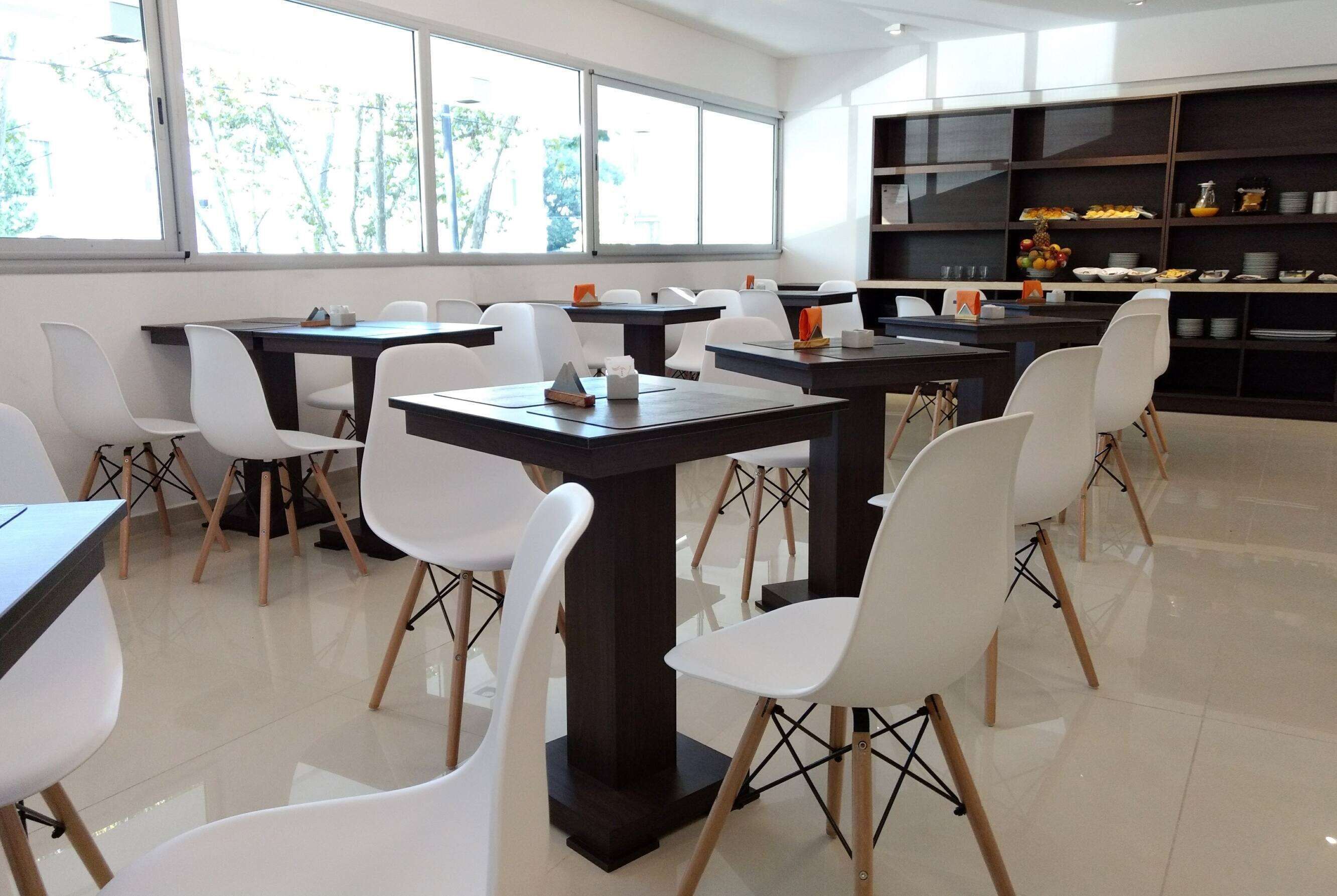 A modern dining area with multiple tables and white chairs, large windows, and a kitchen area with shelves.