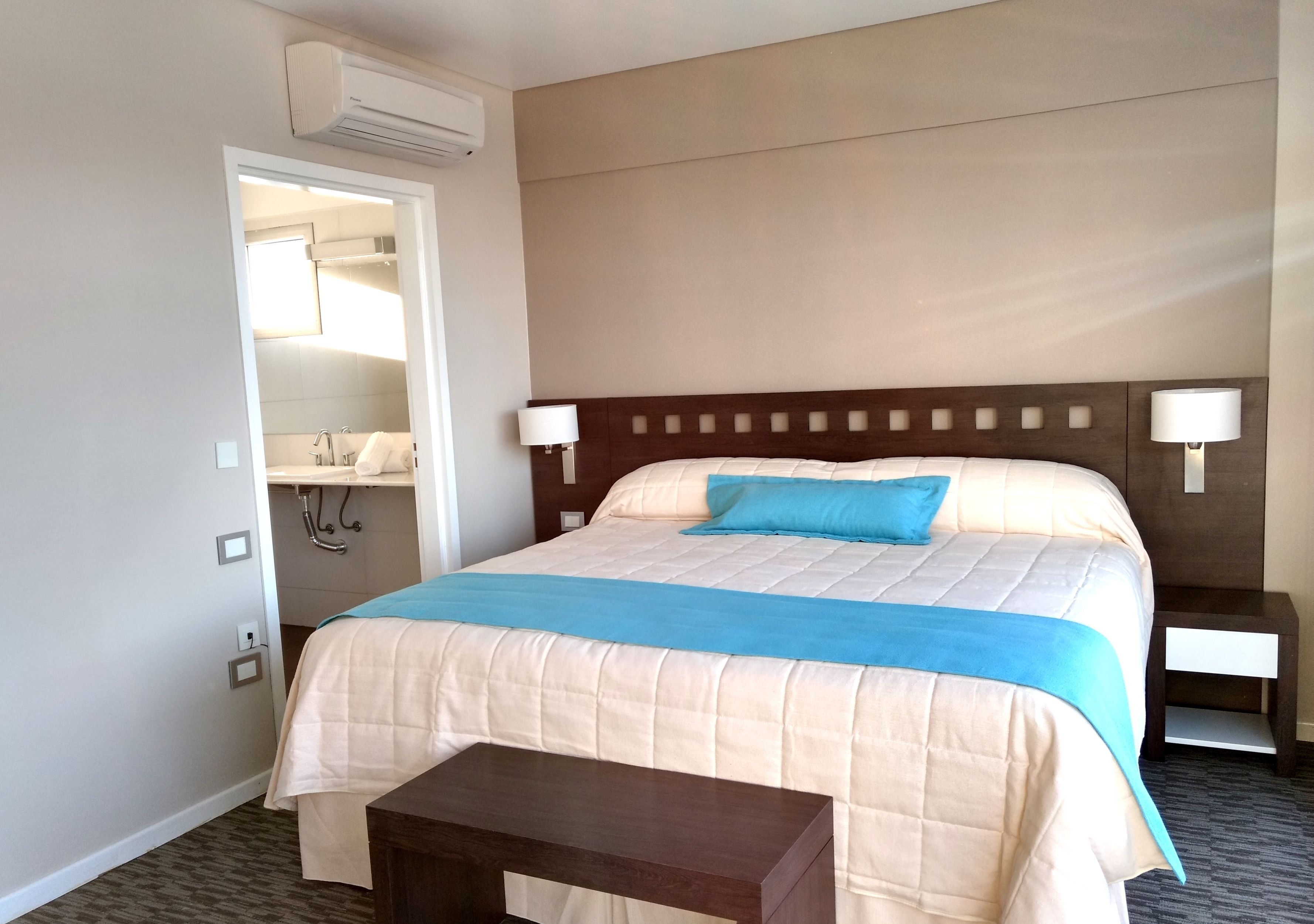 Una habitación de hotel con una cama grande, lámparas de noche, una mesita pequeña y un equipo de aire acondicionado.
