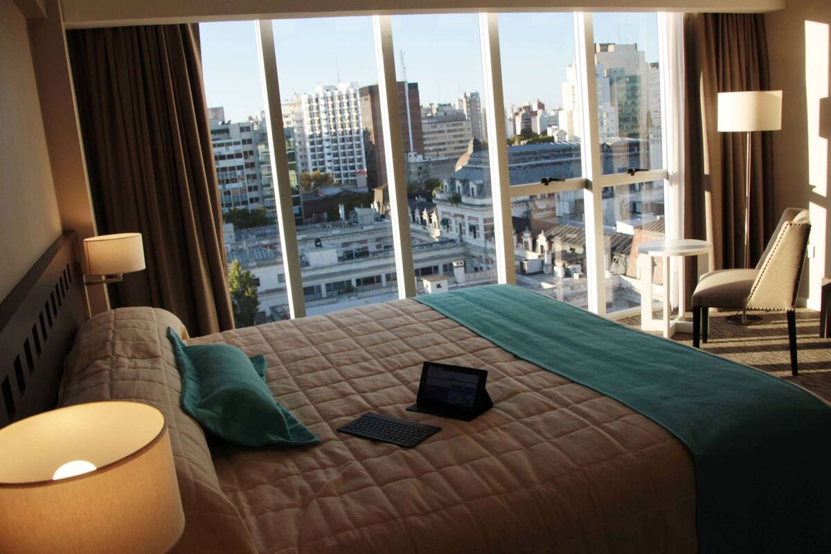 Ein Hotelzimmer mit einem großen Bett, einem Laptop und Blick auf die Stadt durch große Fenster.
