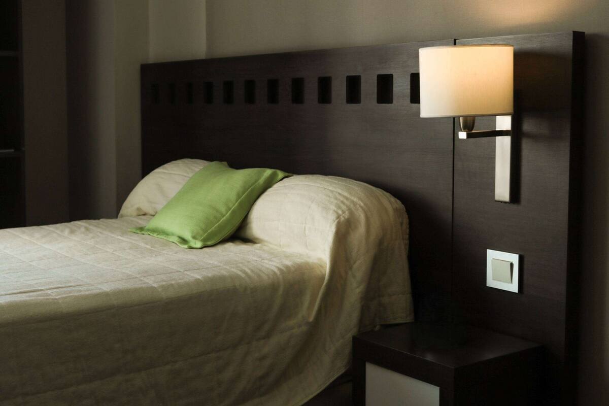 Ein ordentlich bezogenes Bett mit einem grünen Kissen, einem dunklen Kopfteil und einem an der Wand befestigten Nachttisch mit Lampe.