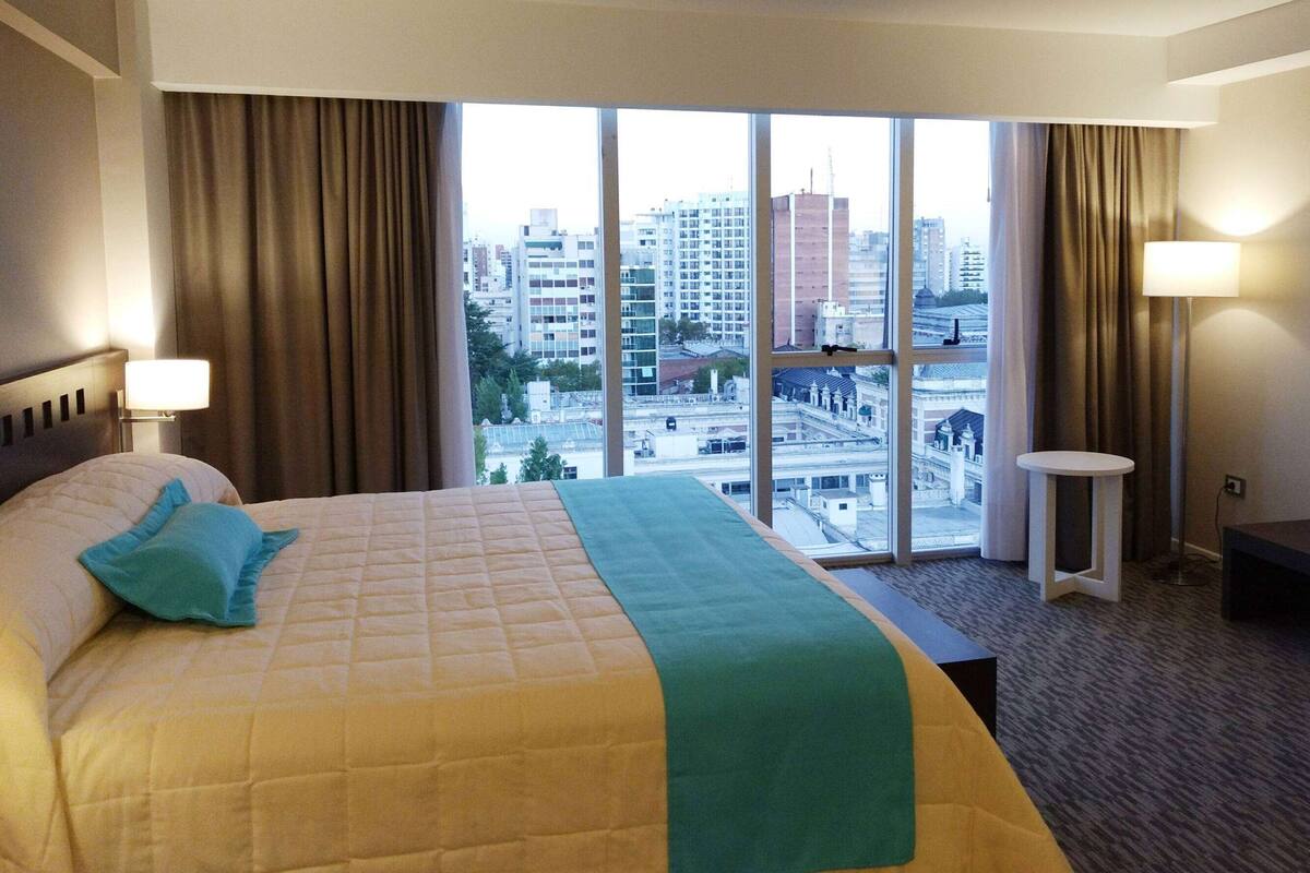Ein Hotelzimmer mit einem großen Bett, Blick auf die Stadt durch ein großes Fenster und einem kleinen Tisch am Fenster.