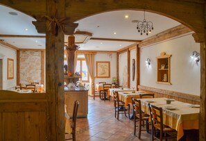 A restaurant with wooden beams, stone walls, and a central island. Tables are set with orange tablecloths and wooden chairs. - Antica Residenza de Poda (Contà)