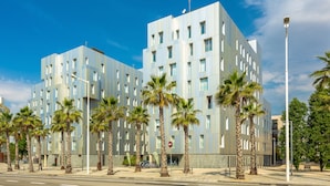 A modern building with a grid-like facade, palm trees in front, and a clear blue sky. - Urban District - Rambla Suites & Pool (Barcelona)