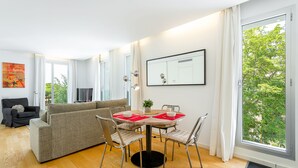 A modern living room with a grey sofa, a dining table with chairs, and a large window with a view of trees. - Urban District - Rambla Suites & Pool (Barcelona)