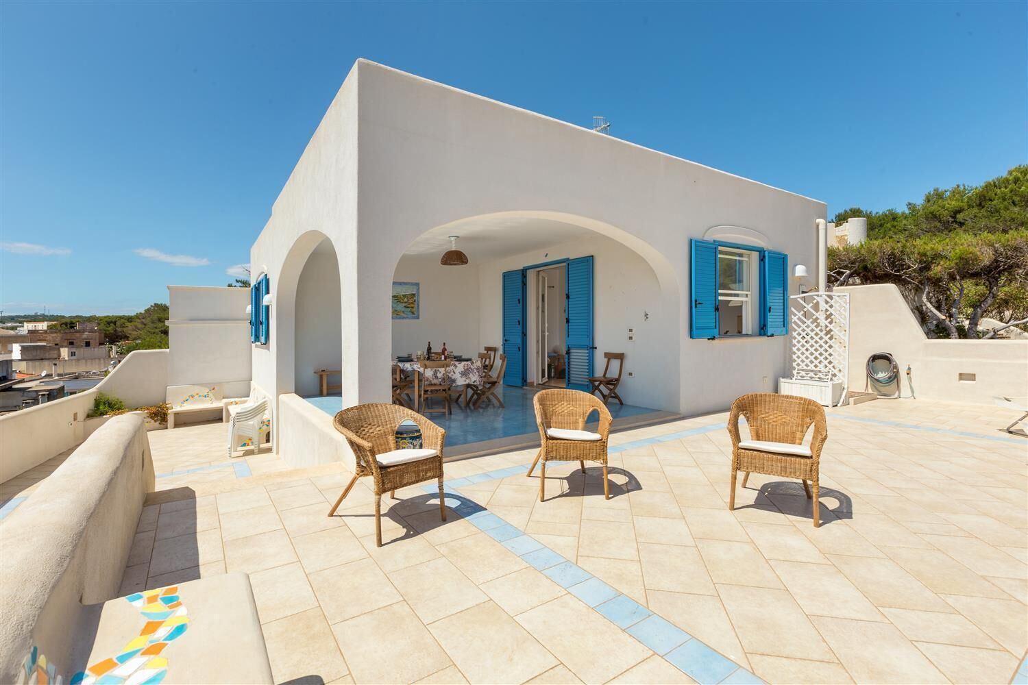 A modern white building with blue shutters, a patio with wicker chairs, and a dining area visible through an archway.