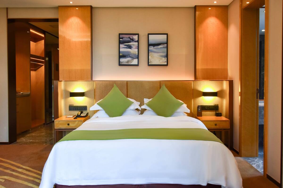 A hotel room with a large bed, green pillows, two bedside tables with lamps, and artwork on the wall.