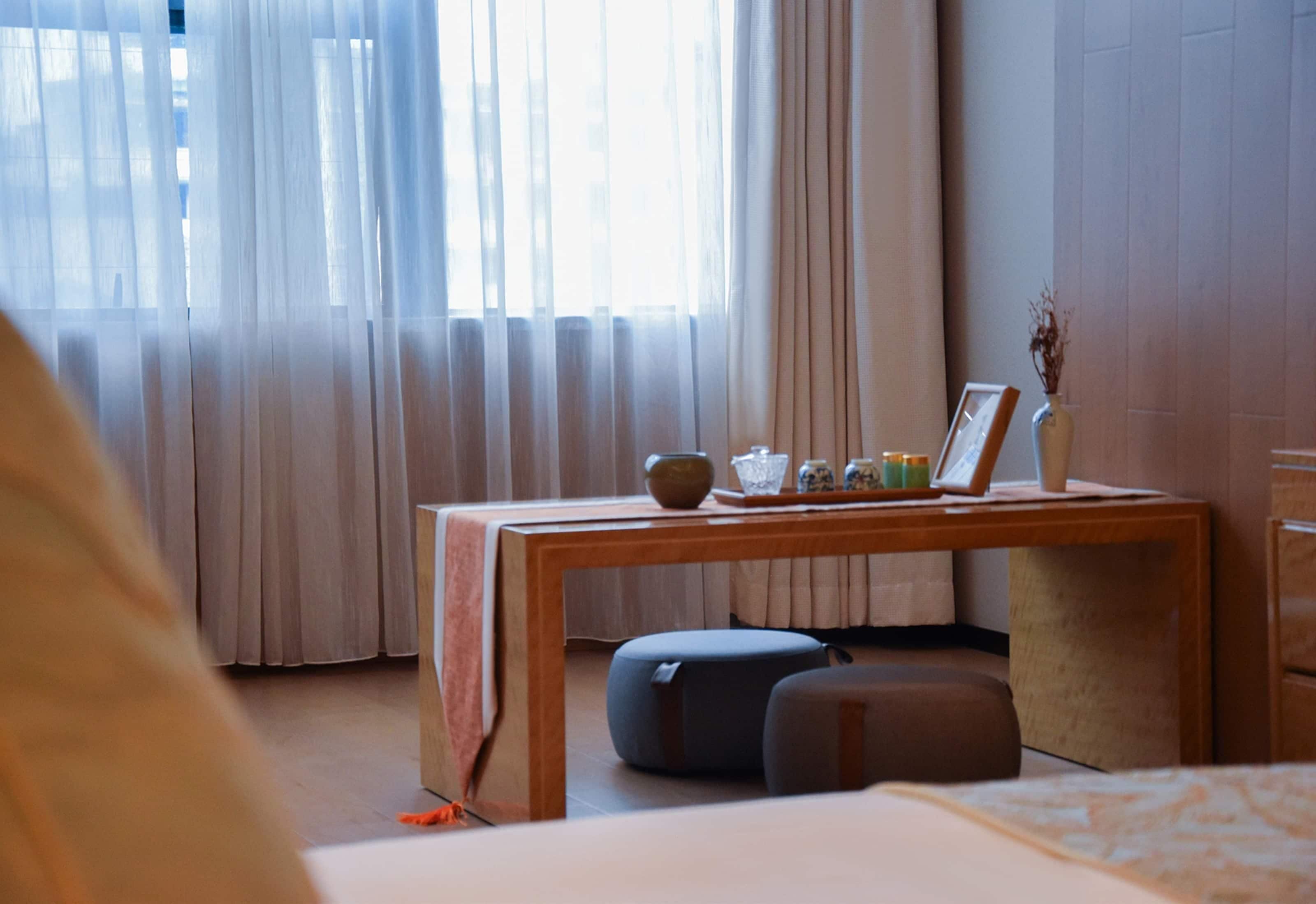 A hotel room with a wooden table, two ottomans, a vase with dried plants, and a window with sheer curtains.