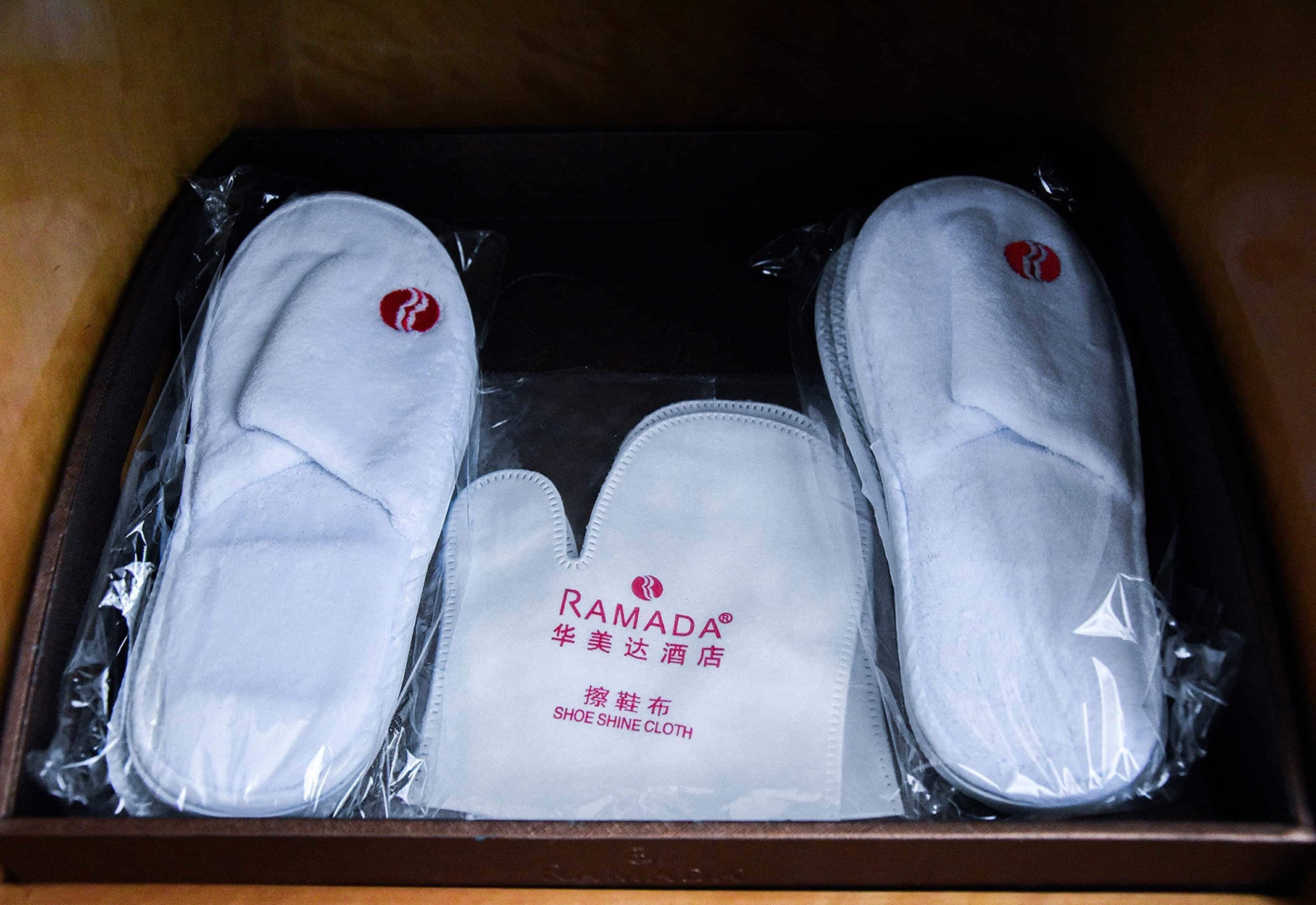 A box containing three pairs of white slippers with a red logo, labeled as "RAMADA" and "SHOE SHINE CLOTH."