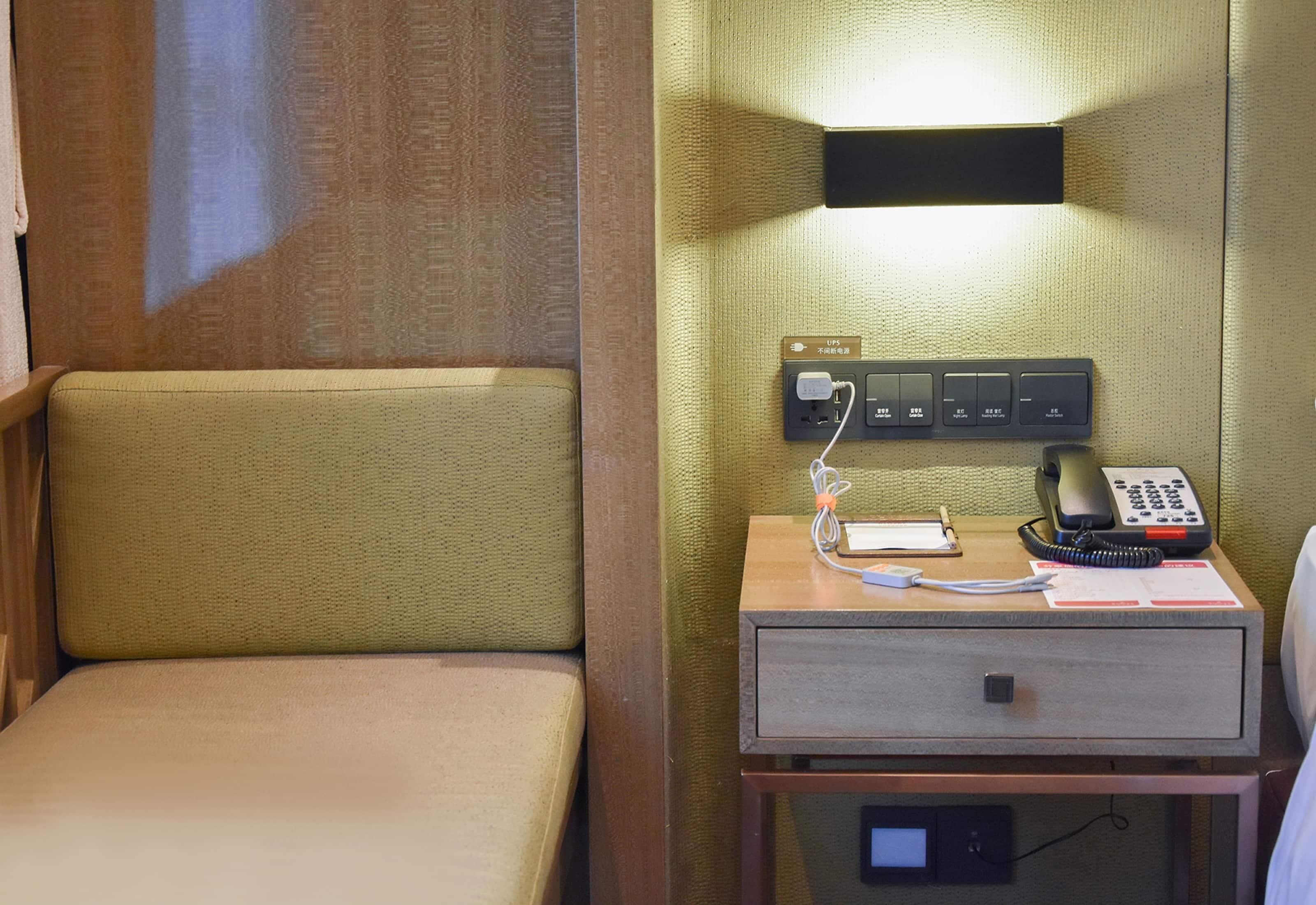 Une chambre d’hôtel avec un lit simple, une table de chevet avec un téléphone et une multiprise, et une lampe fixée au mur.