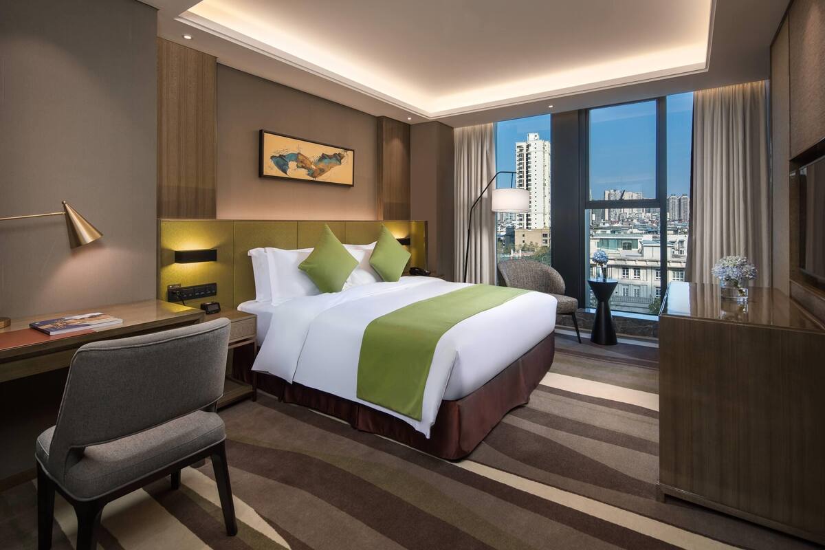 A hotel room with a large bed, a desk, a chair, a desk lamp, a nightstand, a window with a view of the city, and a painting on the wall.