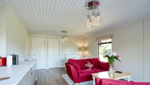 Una sala moderna con un sofá rojo, una mesa de centro blanca y un jarrón con flores. Hay una zona de cocina con encimera blanca y un electrodoméstico rojo. La habitación tiene suelo de madera y techo blanco con luces colgantes.