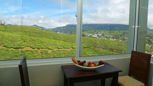 A room with a large window offering a view of a green landscape and a distant town. A table with a fruit bowl is visible in the foreground. - Lourdes Hotel (Nuwara Eliya)
