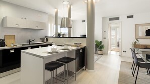 A modern kitchen with a central island, pendant lighting, and a dining area with a glass table and black chairs. - Amazing skyline with amazing views (Barcelona)