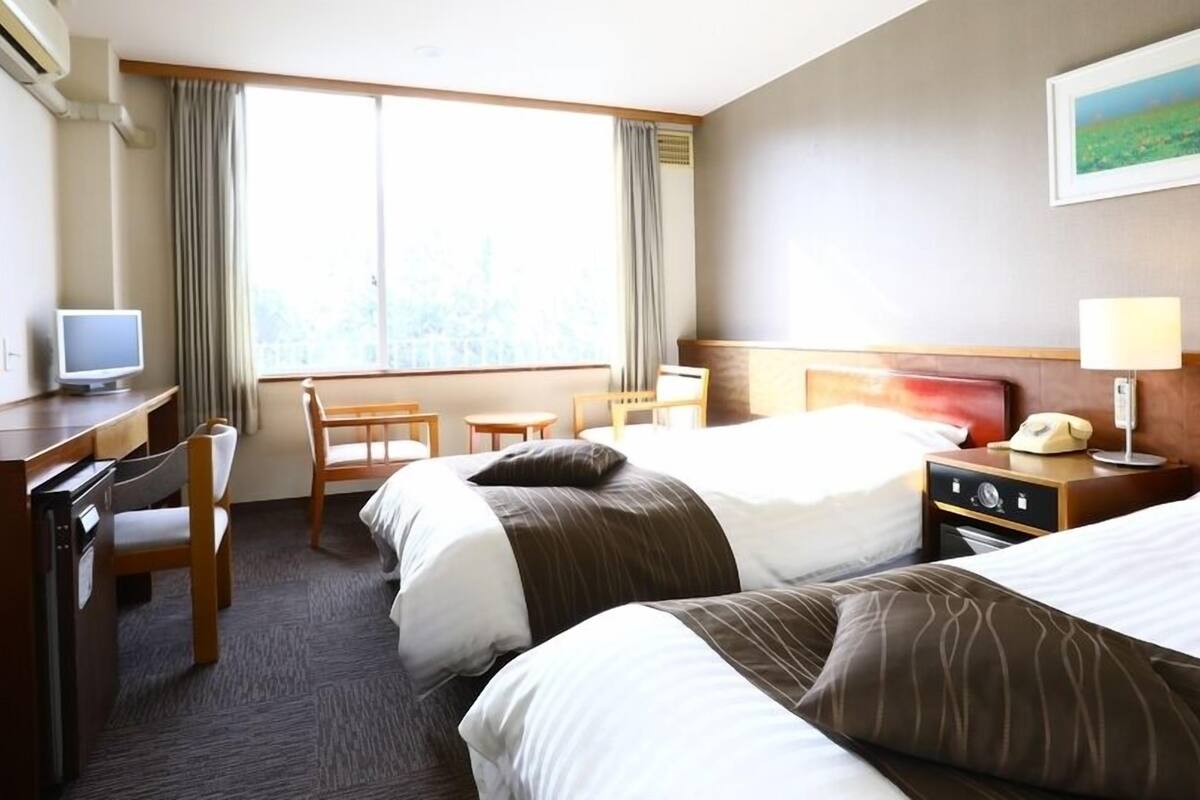 Habitación de hotel con dos camas, un escritorio con computadora, una silla, televisor y ventana con cortinas.