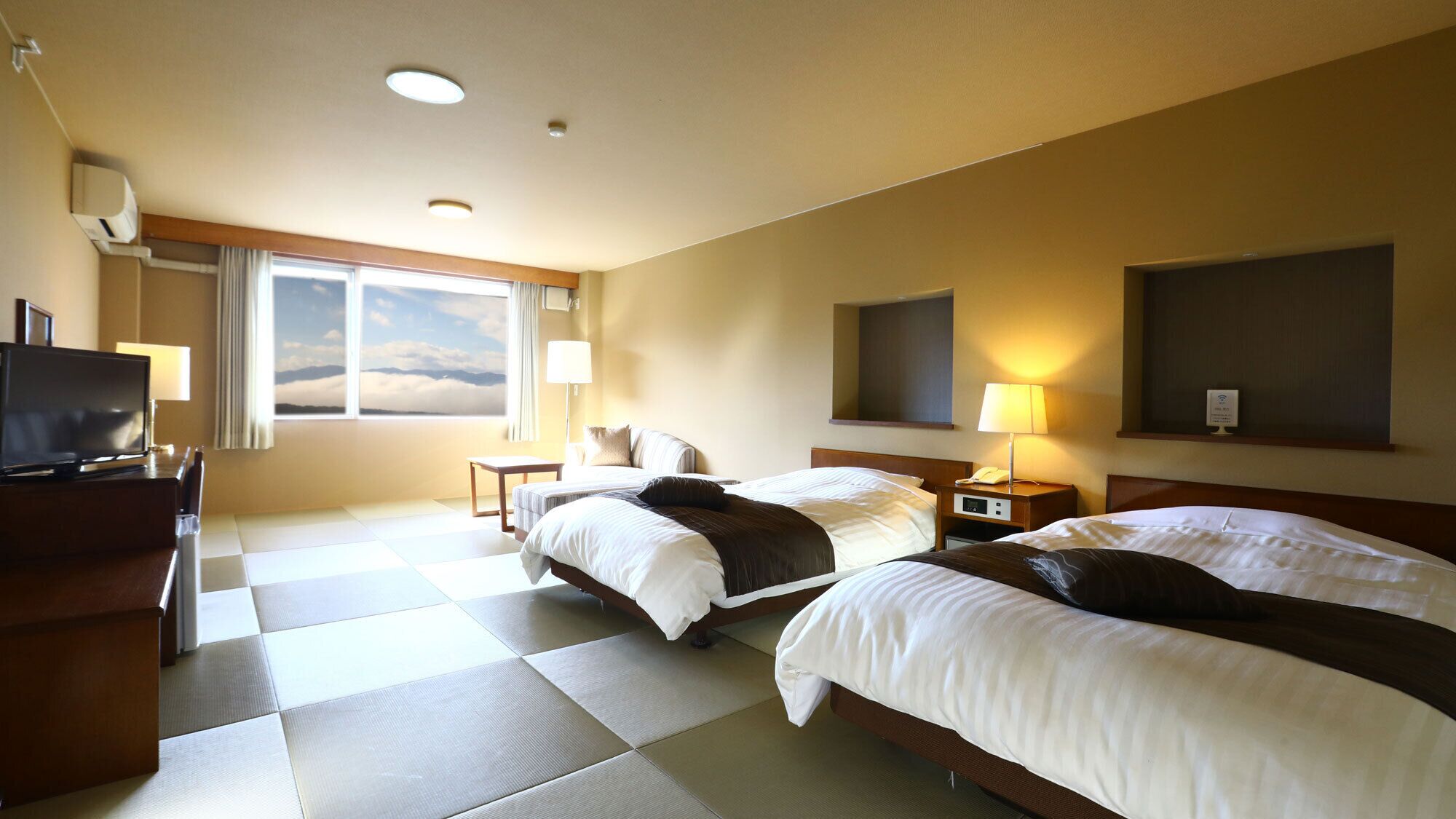 A hotel room with two beds, a TV, a desk, and a window with a view of the sky.