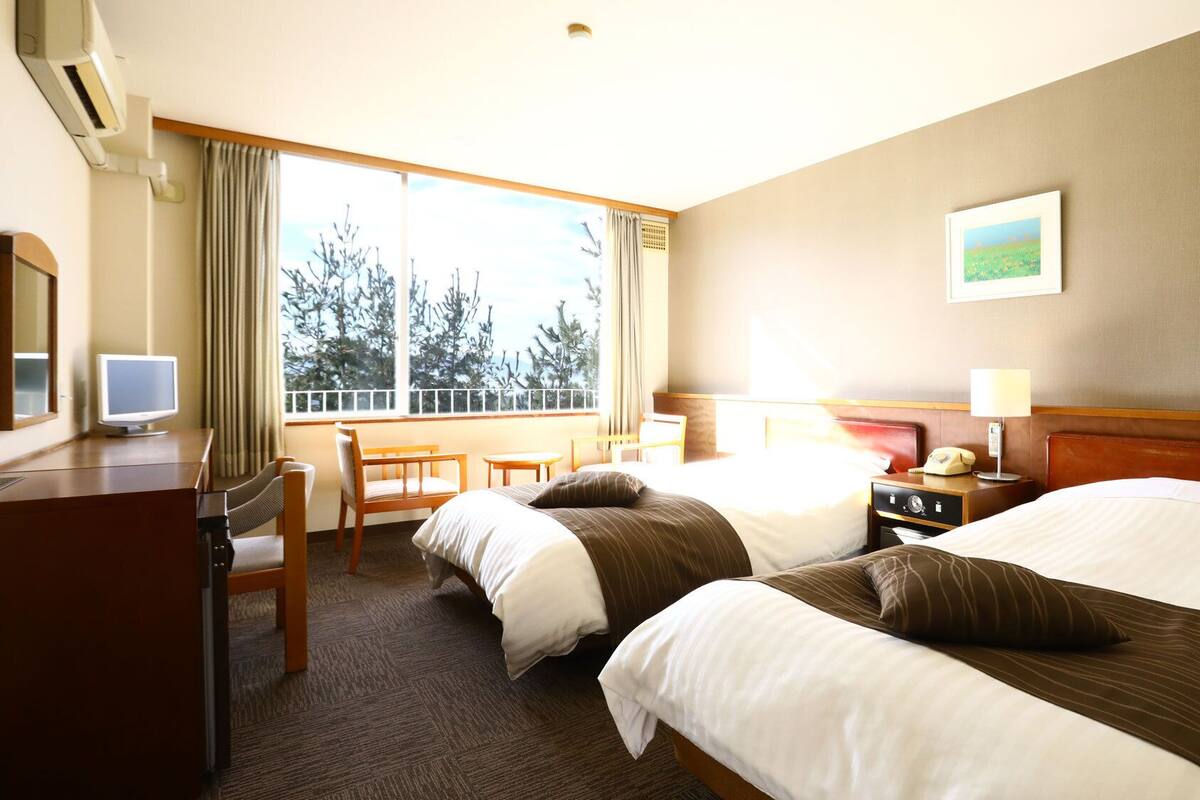 Habitación de hotel con dos camas, un escritorio con computadora, teléfono y ventana con vista a árboles.