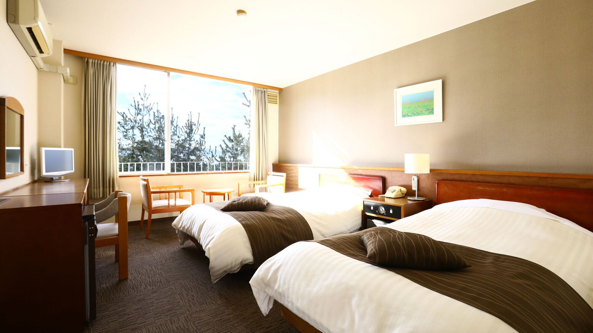 A hotel room with two beds, a desk with a computer, a telephone, and a window with a view of trees.