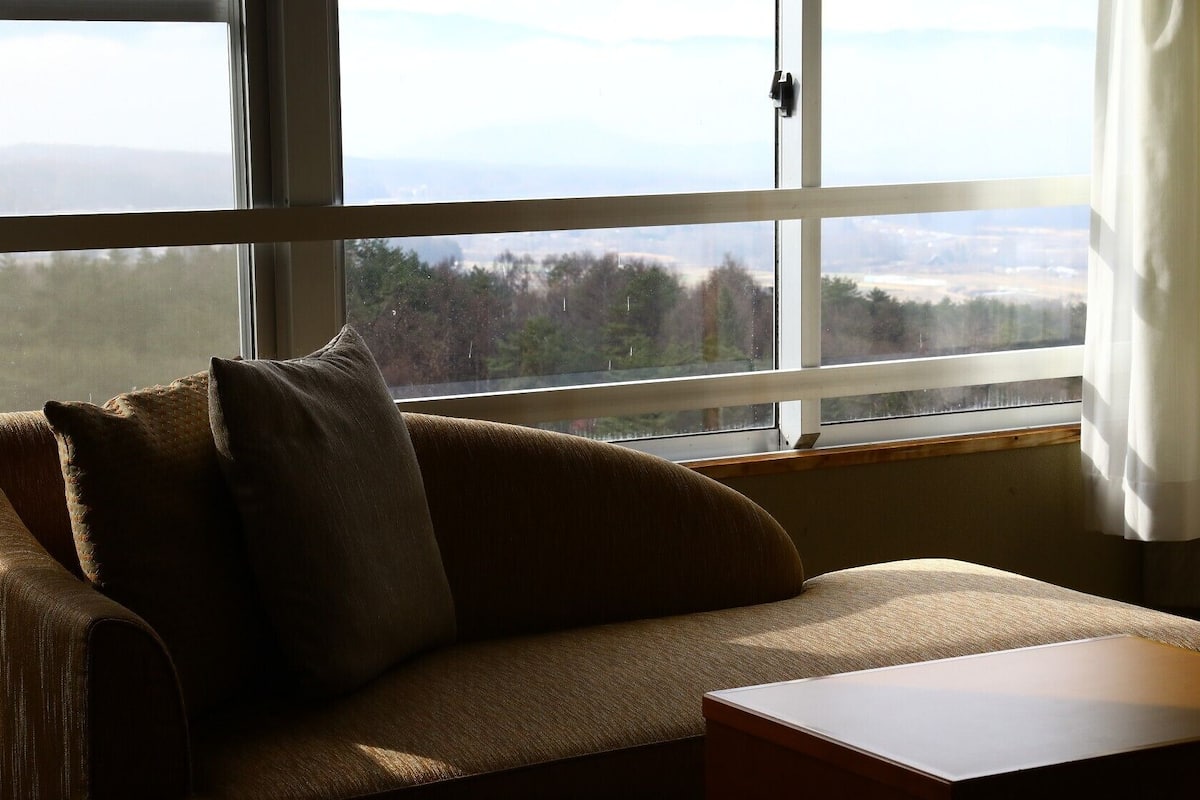 Una habitación con sofá, una mesita y ventana que da a árboles y un paisaje lejano.