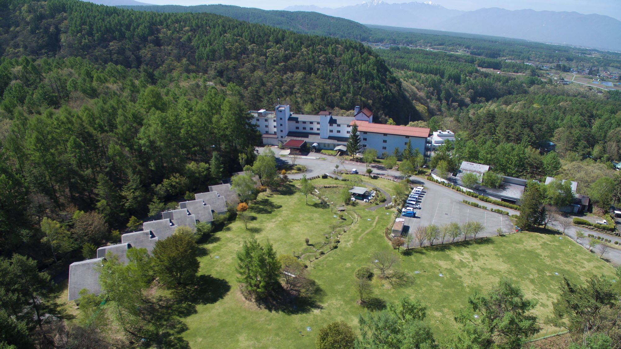 Vista aérea de un hotel con estacionamiento, rodeado de colinas boscosas.