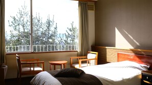 Een hotelkamer met een bed, twee stoelen, een kleintje tafeltje, een raam met uitzicht op bomen, en een gordijn.