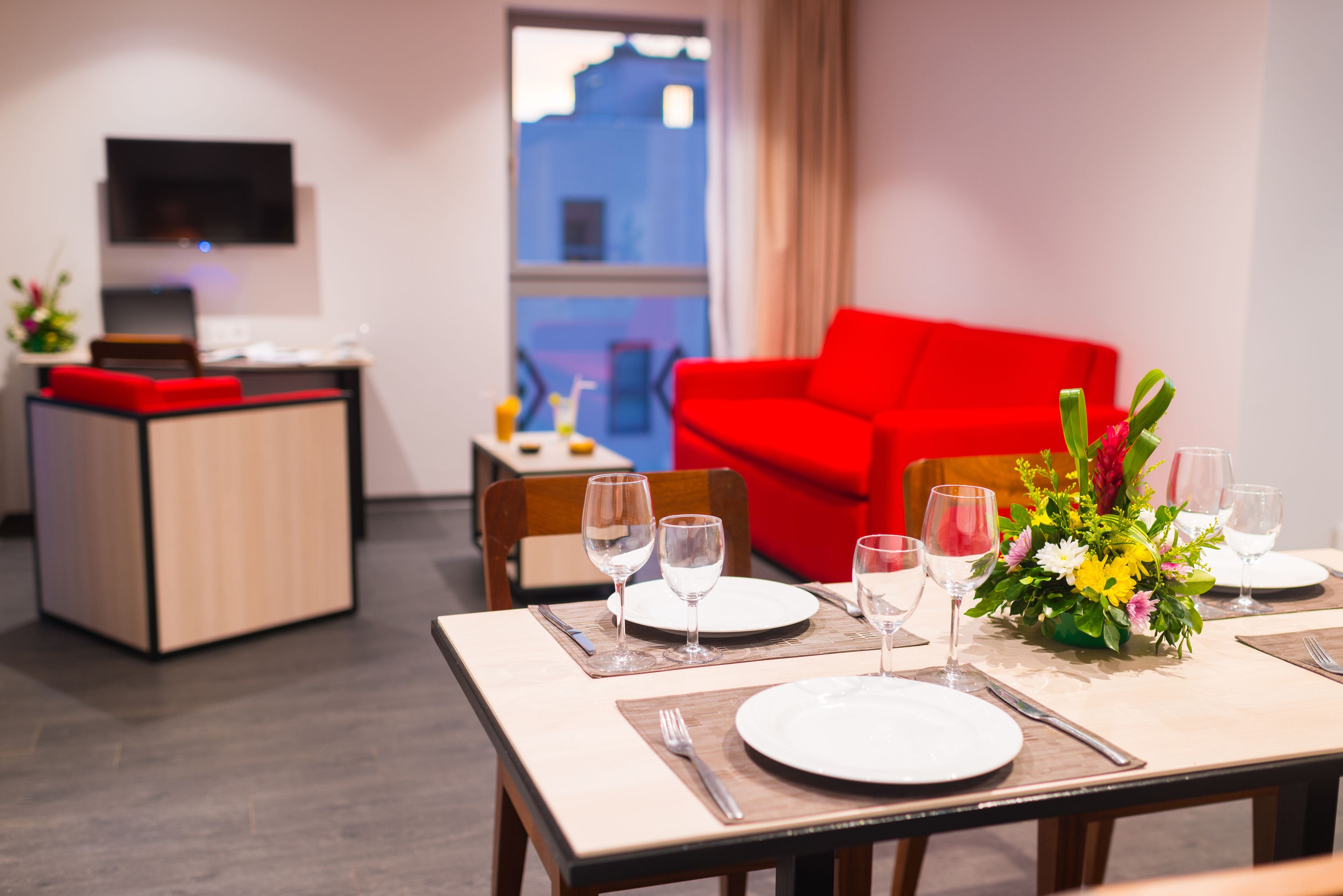 A dining area with a table set for two, a red sofa, and a TV mounted on the wall.