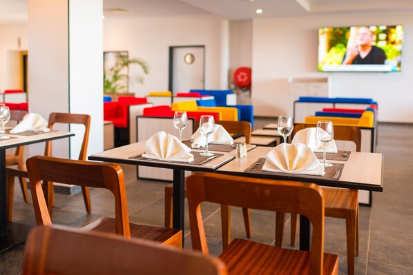 A dining area with tables set for two, each with a folded napkin, a wine glass, and cutlery. There are colorful chairs and a television mounted on the wall in the background.