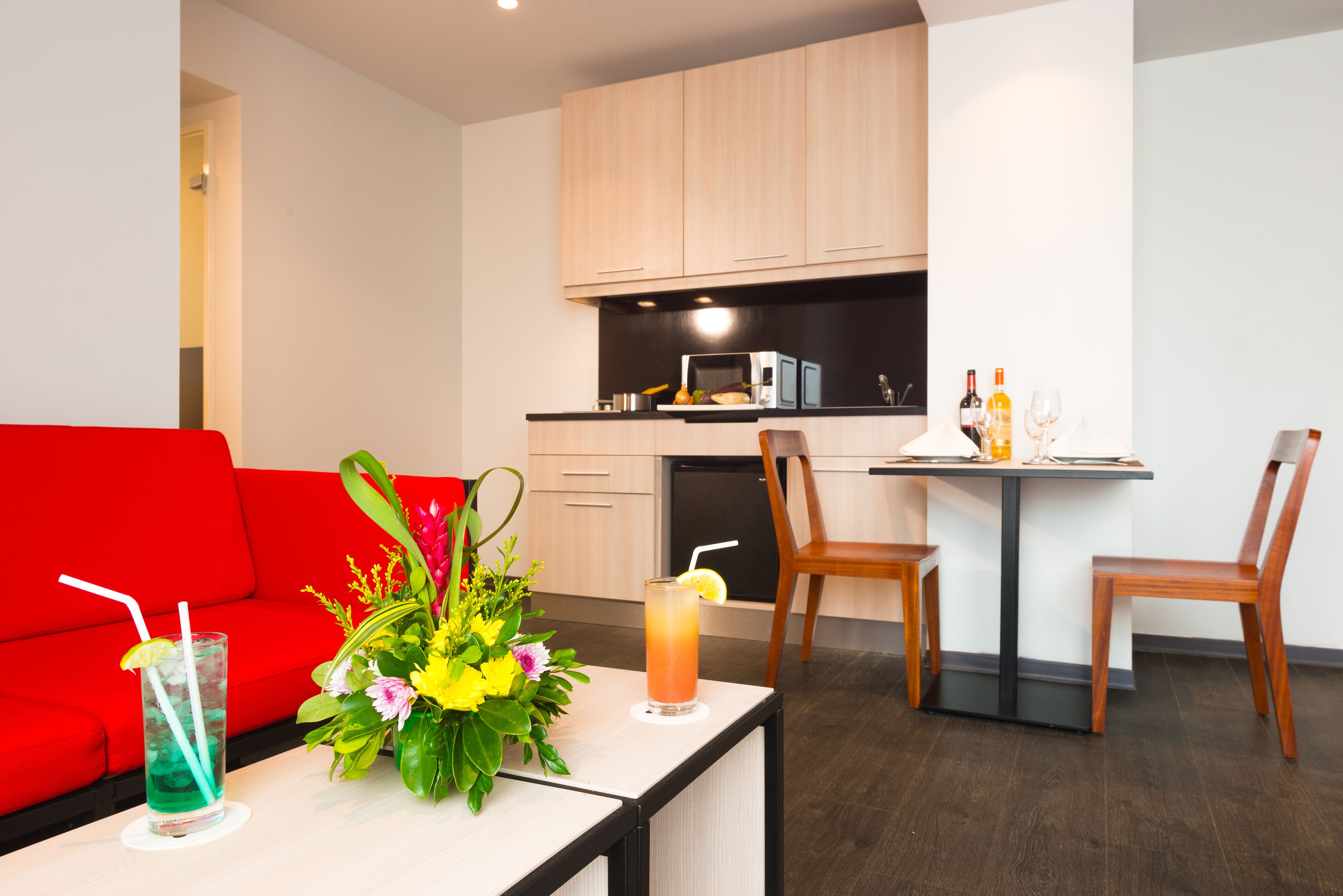A modern kitchen with wooden cabinets, a dining table with two chairs, and a red sofa. There is a glass of iced tea and a floral arrangement on the table.