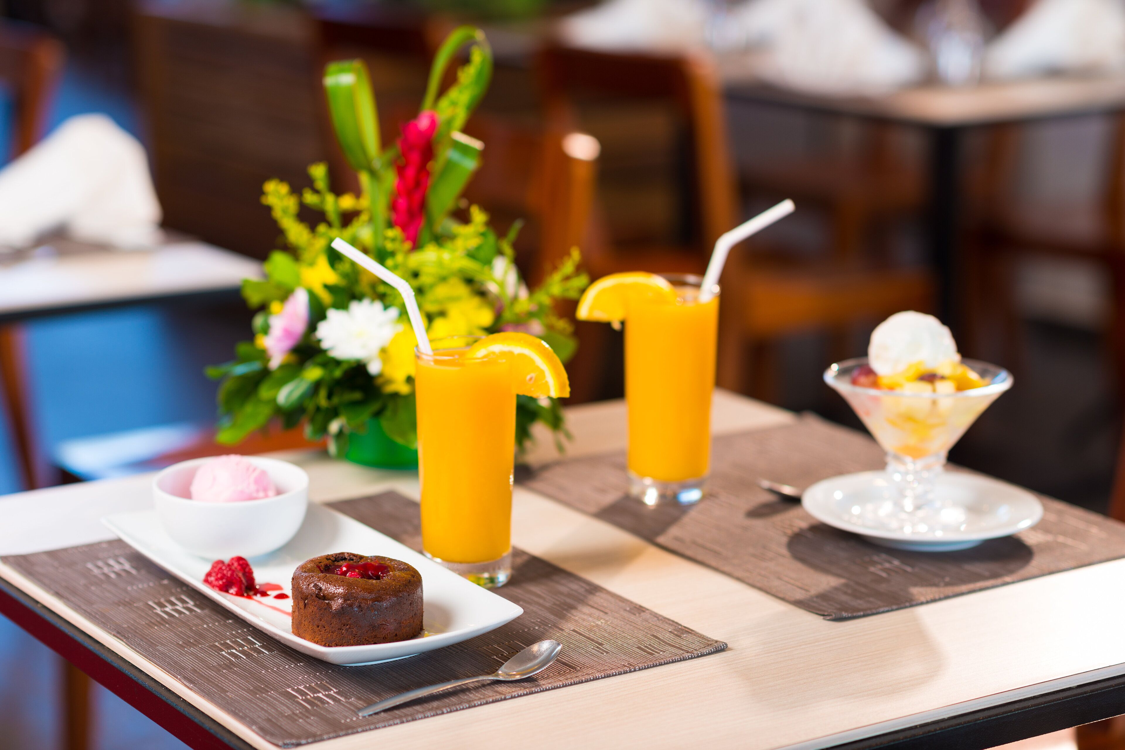 A dining table with a dessert plate, two glasses of orange juice, a bowl of ice cream, and a floral centerpiece.