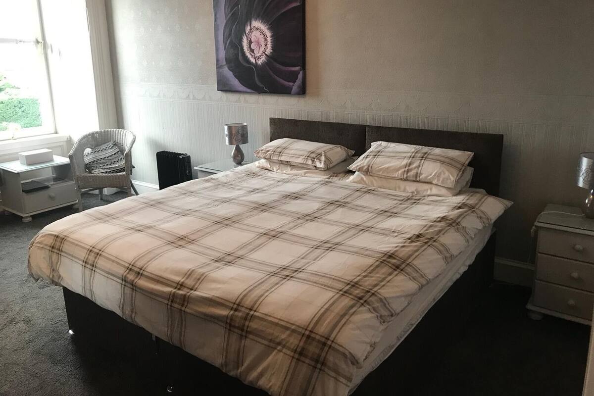 A neatly made bed with a plaid bedspread, a bedside table with a lamp, a chair, and a wall-mounted artwork.