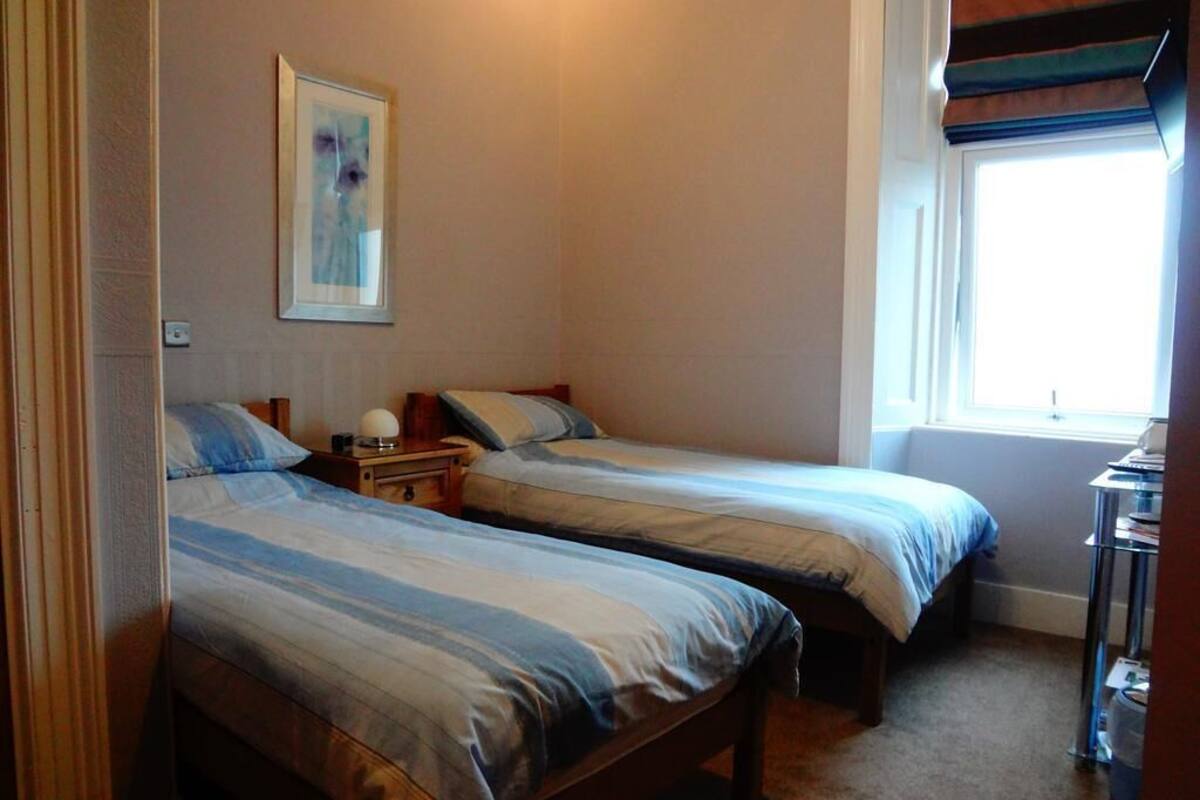 A room with two single beds, a bedside table with a lamp, a framed picture on the wall, and a window with a view.