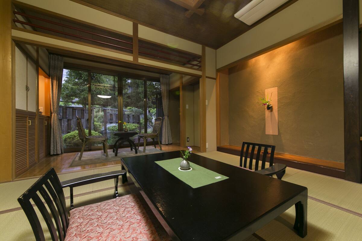 Ein traditioneller japanischer Raum mit Tatami-Boden, einem niedrigen Tisch mit Vase und Schiebetüren zu einem Außenbereich.