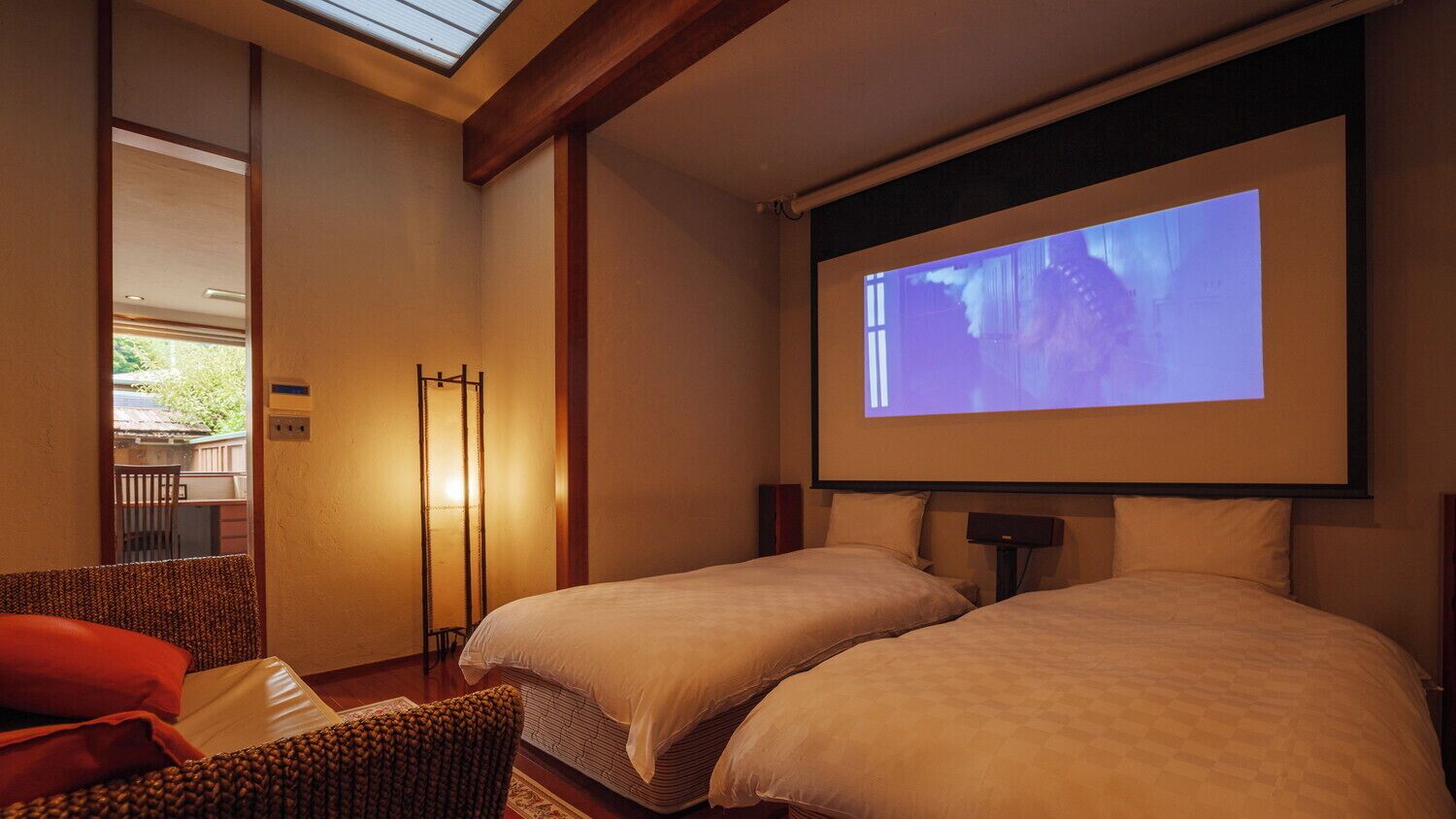 A hotel room with two beds, a projector screen, and a seating area with a chair.