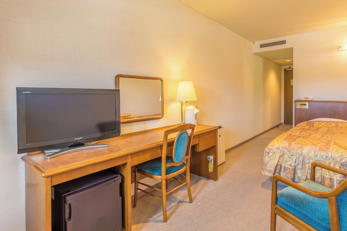 Habitación de hotel con cama, escritorio con televisor, silla, lámpara y refrigerador pequeño.