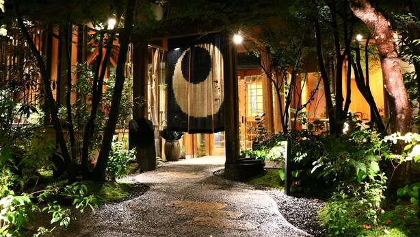A pathway leading to a building with traditional wooden architecture, surrounded by lush greenery and a decorative hanging element. - Tsumikusa no Yado Komatsu (Kirishima)