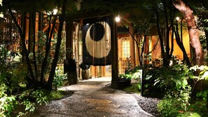 A pathway leading to a building with traditional wooden architecture, surrounded by lush greenery and a decorative hanging element. - Tsumikusa no Yado Komatsu (Kirishima)