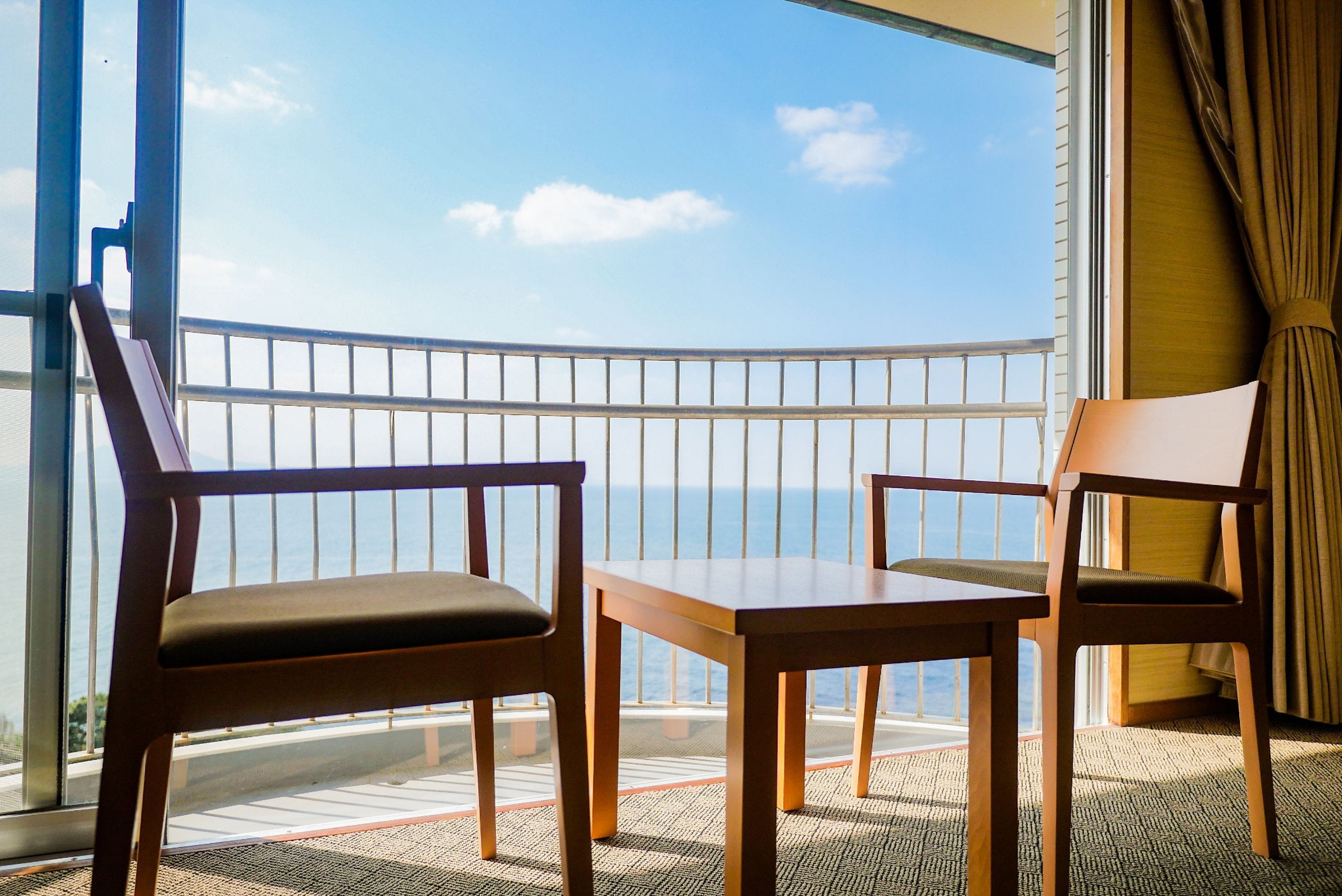 A balcony with two wooden chairs and a table, overlooking a clear blue sky and a distant shoreline.