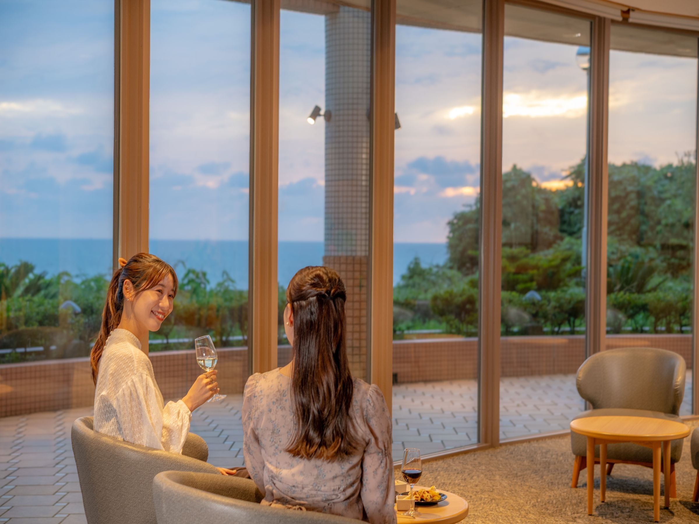 Dos personas sentadas en una terraza con vista al mar, una mesa con un plato y una copa, y una silla.